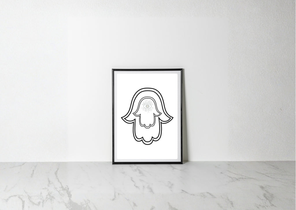 A minimalist black and white art print of a hamsa hand with a skull detail, displayed in a black frame on a light marble surface against a white wall.
