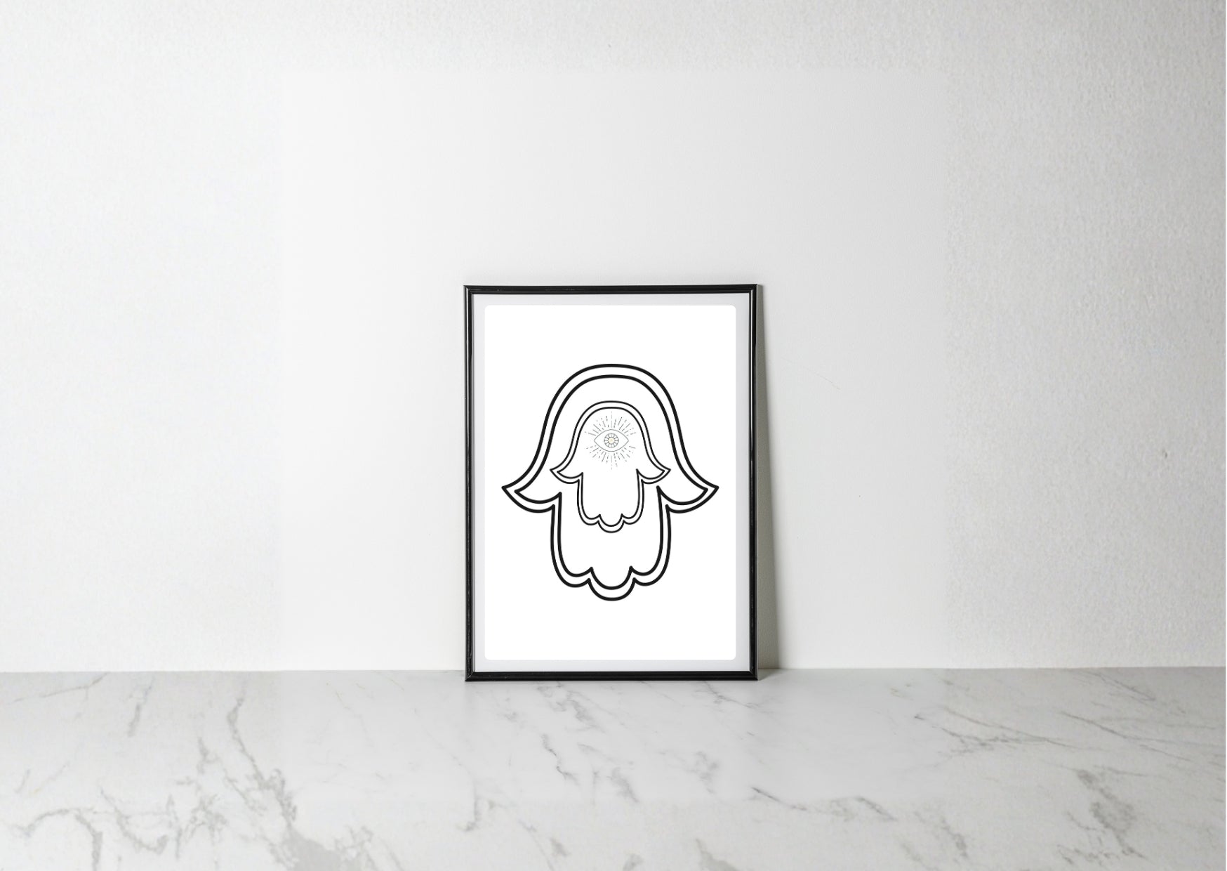 A minimalist black and white art print of a hamsa hand with a skull detail, displayed in a black frame on a light marble surface against a white wall.