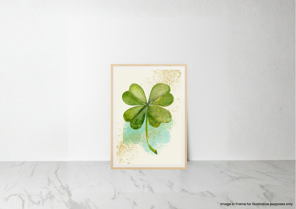 A framed art print featuring a watercolor green four-leaf clover on a cream background with gold glitter accents and a light blue wash, leaning against a white wall on a marble surface.