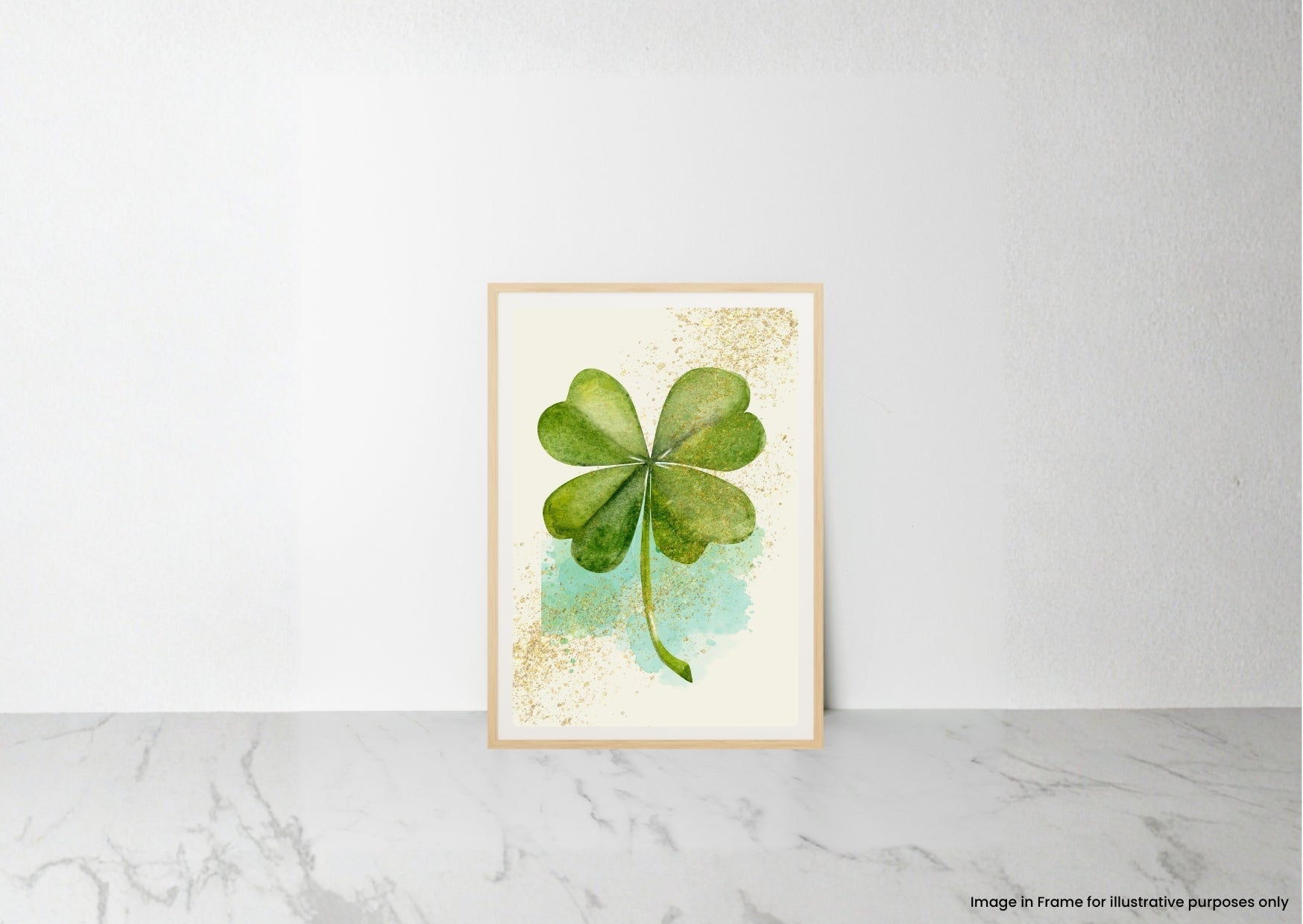 A framed art print featuring a watercolor green four-leaf clover on a cream background with gold glitter accents and a light blue wash, leaning against a white wall on a marble surface.