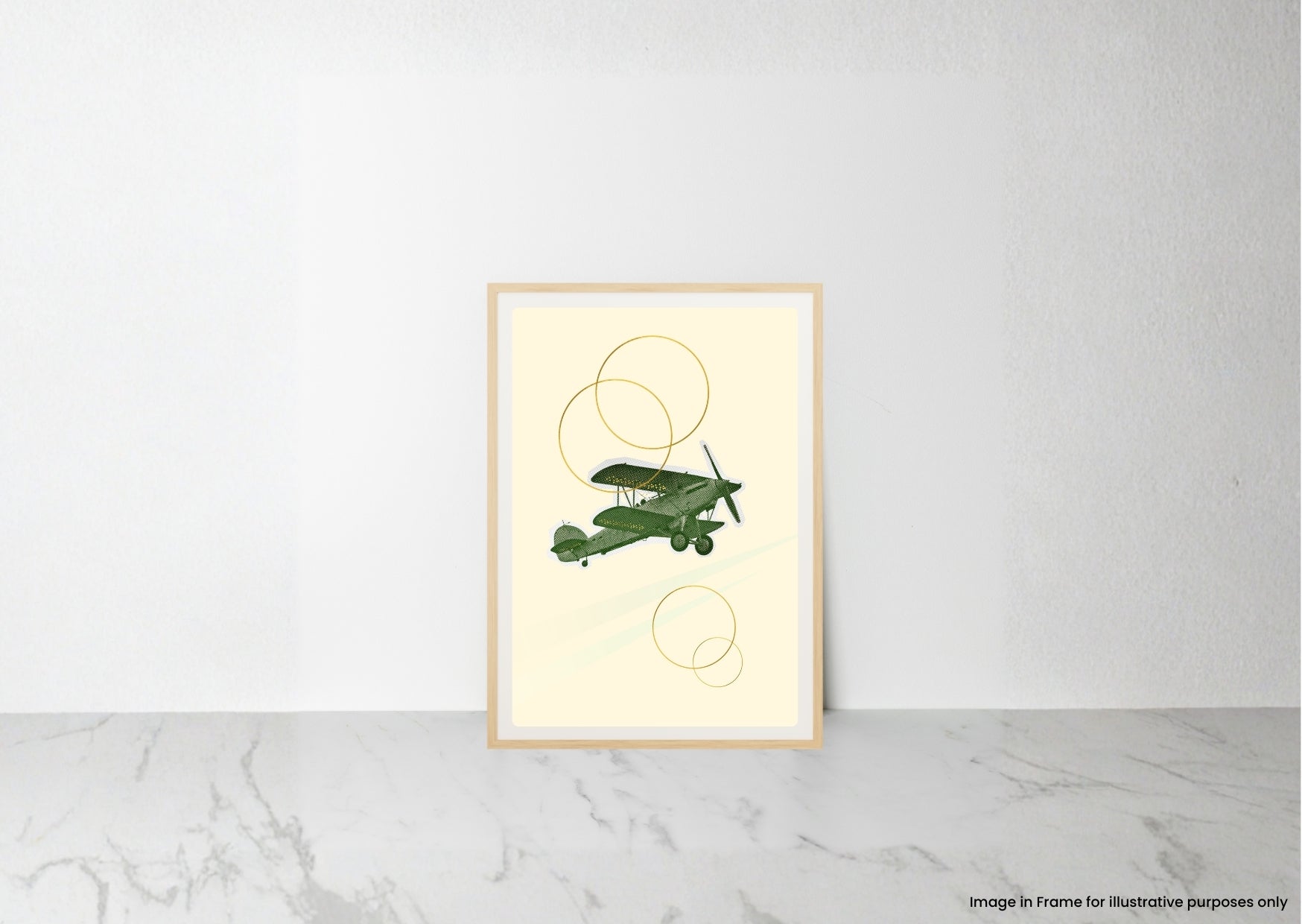 A framed art print featuring a green vintage biplane illustration with gold abstract circles on a cream background, leaning against a white wall on a light marble surface.