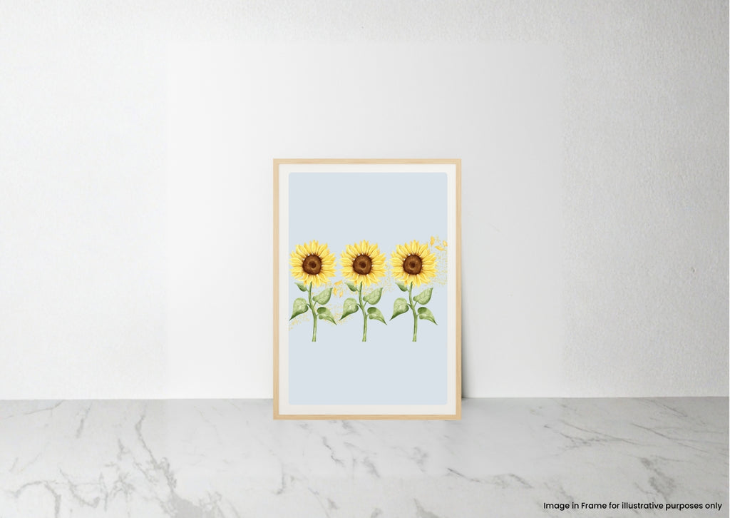 A vertical framed art print featuring three watercolor sunflowers on a pale blue background, leaning against a white wall on a light marble surface.